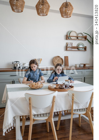 Two girls sitting at round kitchen table with notepads and eating pastries together in cozy home. Notepads, table, eating, pastries, kitchen, girls. 141413820