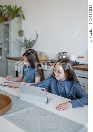 Two girls in denim shirts doing homework at kitchen table with natural daylight. Homework, kitchen, denim, girls, daylight. Two girls in denim shirts doing homework at kitchen table with natural daylight. Homework, kitchen, denim, girls, daylight. 141413853