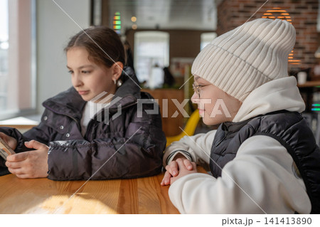 Two girls in winter hats looking at phone and talking at cafe table in warm light. Phone, winter hats, cafe, talking, girls. Two girls in winter hats looking at phone and talking at cafe table in warm light. Phone, winter hats, cafe, talking, girls. 141413890