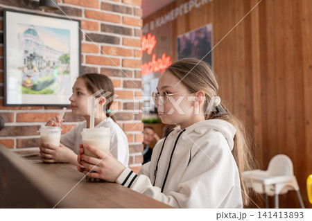 Girls in white sweatshirts drinking milk tea and laughing together at cafe counter in sunlight. Milk tea, cafe, girls, white, laughing. Girls in white sweatshirts drinking milk tea and laughing together at cafe counter in sunlight. Milk tea, cafe, girls, white, laughing. 141413893
