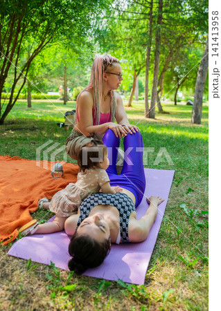 Yoga instructor working one-on-one with mother and child in assisted abs exercise on mat in park. Assisted, abs, yoga, instructor, mat, mother, child, park. Yoga instructor working one-on-one with mother and child in assisted abs exercise on mat in park. Assisted, abs, yoga, instructor, mat, mother, child, park. 141413958