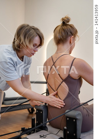 A woman in activewear does pilates exercises for the chest and back muscles with a reformer machine in a sunlite studio, committed to her workout routine A woman in activewear does pilates exercises for the chest and back muscles with a reformer machine in a sunlite studio, committed to her workout routine 141413959