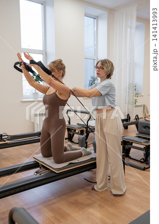 A woman in activewear does pilates exercises for the chest and back muscles with a reformer machine in a sunlite studio, committed to her workout routine A woman in activewear does pilates exercises for the chest and back muscles with a reformer machine in a sunlite studio, committed to her workout routine 141413993