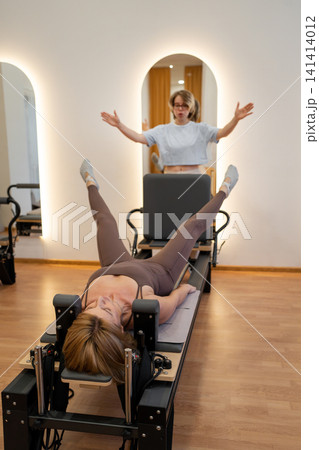 Female instructor guiding client through leg extension routine on pilates reformer in airy gym. Leg extension, pilates reformer, instructor, routine, gym Female instructor guiding client through leg extension routine on pilates reformer in airy gym. Leg extension, pilates reformer, instructor, routine, gym 141414012
