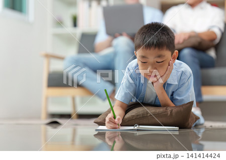 Asian boy doing homework on floor with parents working in background Asian boy doing homework on floor with parents working in background 141414424