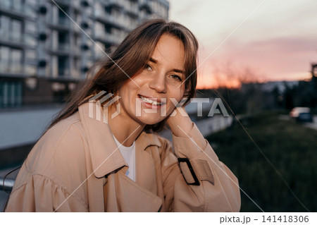 Woman sunset trench coat smiling outdoors against a city apartment backdrop during golden hour in a joyful mood Woman sunset trench coat smiling outdoors against a city apartment backdrop during golden hour in a joyful mood 141418306