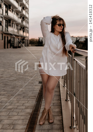 Woman fashion model posing in white blouse and stockings on an urban balcony at sunset with modern buildings Woman fashion model posing in white blouse and stockings on an urban balcony at sunset with modern buildings 141418312