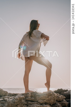 Woman beach sunset standing on rocky ocean shore in white shirt during golden hour enjoying summer vacation and peace Woman beach sunset standing on rocky ocean shore in white shirt during golden hour enjoying summer vacation and peace 141418341