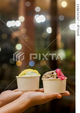 Premium Assorted Artisan Gelato in Paper Cups Held by Hand with Cafe Lights Premium Assorted Artisan Gelato in Paper Cups Held by Hand with Cafe Lights 141419641