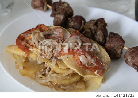 Delicious baked meat skewer served on a white plate with potato casserole, tomatoes and onions, ready for a tasty homemade lunch or dinner meal, top view close up. Delicious baked meat skewer served on a white plate with potato casserole, tomatoes and onions, ready for a tasty homemade lunch or dinner meal, top view close up. 141421823