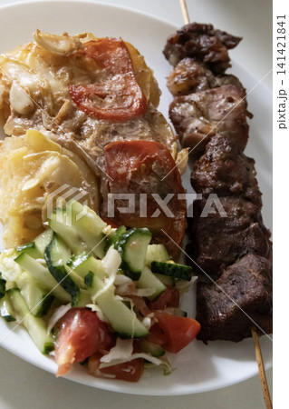 Delicious homemade dinner on a white plate: grilled meat skewer, baked potato casserole with tomatoes, and fresh cucumber and tomato salad. Delicious homemade dinner on a white plate: grilled meat skewer, baked potato casserole with tomatoes, and fresh cucumber and tomato salad. 141421841