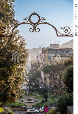 Naples, italy. December 30, 2024. Capodimonte park view of naples cityscape, seeing buildings, treelined street, and distant dome framed by an ornate iron arch 141422043