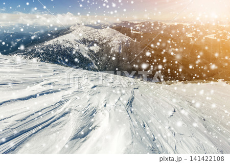 Beautiful winter landscape. Steep mountain hill slope with white deep snow, distant mountain range panorama, large snowflakes and bright shining sun rays on blue sky colorful copy space background. Beautiful winter landscape. Steep mountain hill slope with white deep snow, distant mountain range panorama, large snowflakes and bright shining sun rays on blue sky colorful copy space background. 141422108
