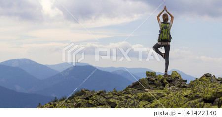 Back view of young slim tourist girl with backpack standing on one leg in yoga pose on rocky top on bright blue morning sky and foggy mountains background. Tourism, traveling and climbing concept. Back view of young slim tourist girl with backpack standing on one leg in yoga pose on rocky top on bright blue morning sky and foggy mountains background. Tourism, traveling and climbing concept. 141422130