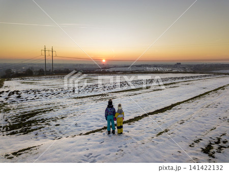 Back view of two young children in warm clothing standing in frozen snow field holding hands on copy space background of setting sun and clear blue sky. Back view of two young children in warm clothing standing in frozen snow field holding hands on copy space background of setting sun and clear blue sky. 141422132