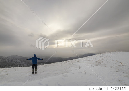 Back view of tourist hiker in warm clothing with backpack standing with raised arms on clearing covered with snow on spruce forest mountain and cloudy sky copy space background. Back view of tourist hiker in warm clothing with backpack standing with raised arms on clearing covered with snow on spruce forest mountain and cloudy sky copy space background. 141422135