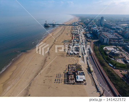 Beautiful high altitude aerial panoramic perspective of a long sandy beach resort promenade with beach clubs and iconic pier with ferris wheel along the North Sea coast under a blue sky Beautiful high altitude aerial panoramic perspective of a long sandy beach resort promenade with beach clubs and iconic pier with ferris wheel along the North Sea coast under a blue sky 141422572