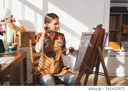 Picture of serious concentrated young Caucasian female artist sitting at desk with painting accessories, holding tube of oil paint, mixing colors on palette; unfinished painting on canvas near her Picture of serious concentrated young Caucasian female artist sitting at desk with painting accessories, holding tube of oil paint, mixing colors on palette; unfinished painting on canvas near her 141422971