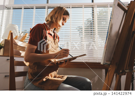 Picture of serious concentrated young Caucasian female artist sitting at desk with painting accessories, holding tube of oil paint, mixing colors on palette; unfinished painting on canvas near her 141422974