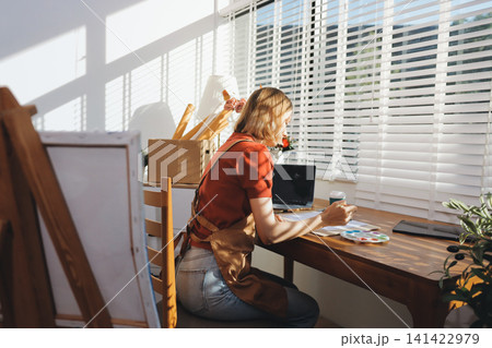 Picture of serious concentrated young Caucasian female artist sitting at desk with painting accessories, holding tube of oil paint, mixing colors on palette; unfinished painting on canvas near her Picture of serious concentrated young Caucasian female artist sitting at desk with painting accessories, holding tube of oil paint, mixing colors on palette; unfinished painting on canvas near her 141422979