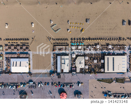 Beautiful top down high altitude aerial panoramic perspective of beach resort clubs with sunbeds umbrellas and colorful vintage cars parked on a paved boulevard promenade under bright sunlight Beautiful top down high altitude aerial panoramic perspective of beach resort clubs with sunbeds umbrellas and colorful vintage cars parked on a paved boulevard promenade under bright sunlight 141423148
