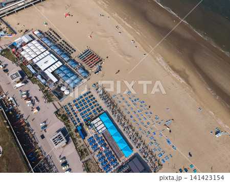 Beautiful high altitude aerial panoramic perspective of beach resort clubs with blue umbrellas lounge chairs and parking near sandy coast line under bright sunlight Beautiful high altitude aerial panoramic perspective of beach resort clubs with blue umbrellas lounge chairs and parking near sandy coast line under bright sunlight 141423154