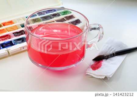 Workspace of a watercolor artist with glass of water, a brush, a stained napkin, and a palette with watercolor paints on a white table. 141423212