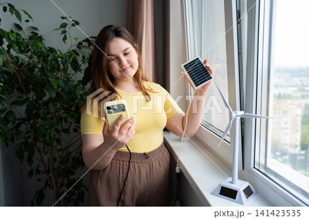 Young woman using smartphone and holding solar panel near window with model wind turbine. Renewable energy technology and smart energy monitoring app 141423535