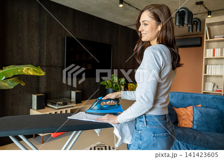 Long-haired young woman standing near the ironing board and ironing laundry Long-haired young woman standing near the ironing board and ironing laundry 141423605