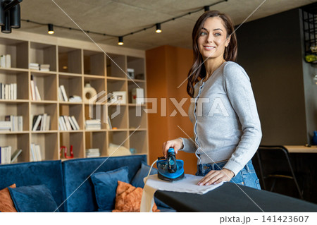 Long-haired young woman standing near the ironing board and ironing laundry 141423607