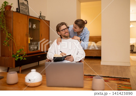 Young woman hugging her husband who is sitting in the arm-chair and making notes Young woman hugging her husband who is sitting in the arm-chair and making notes 141423890