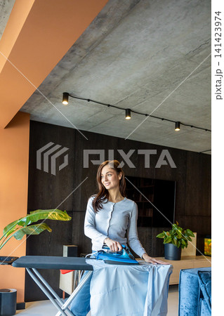 Long-haired young woman standing near the ironing board and ironing laundry Long-haired young woman standing near the ironing board and ironing laundry 141423974