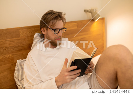 Fair-haired young man sitting in bed with a book in hands Fair-haired young man sitting in bed with a book in hands 141424448
