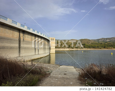 The large concrete arch dam wall of the Beniarres reservoir stretches along the blue water. The area shows land and vegetation near the shoreline. The large concrete arch dam wall of the Beniarres reservoir stretches along the blue water. The area shows land and vegetation near the shoreline. 141424782