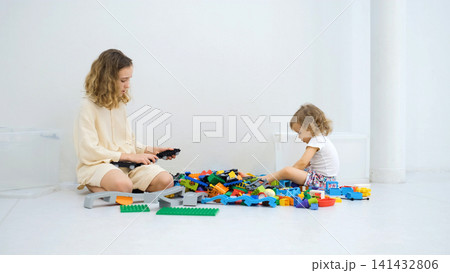 Teen girl sitting beside her little sister helping her sort and organize a pile of colorful construction blocks on the floor 141432806
