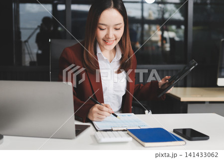 Smiling asian businesswoman talking on phone and using laptop while working at office desk Smiling asian businesswoman talking on phone and using laptop while working at office desk 141433062