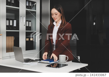 Smiling asian businesswoman talking on phone and using laptop while working at office desk 141433067