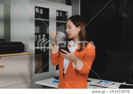 Smiling asian businesswoman talking on phone and using laptop while working at office desk Smiling asian businesswoman talking on phone and using laptop while working at office desk 141433076
