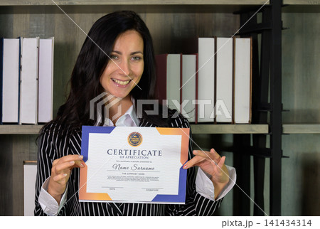 Smiling businesswoman holding certificate of achievement in modern office for professional success recognition and corporate award ceremony concept 141434314