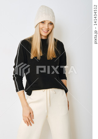 Woman wearing black sweater and white pants standing against light background Woman wearing black sweater and white pants standing against light background 141434512