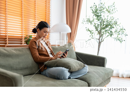 Happy young asian woman relax on comfortable couch at home texting messaging on smartphone 141434550