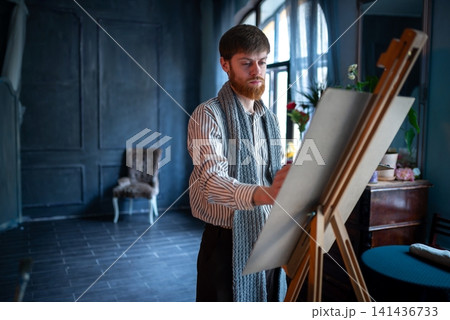 Male painter sketching on large easel in moody studio with wooden furniture and natural window light Male painter sketching on large easel in moody studio with wooden furniture and natural window light 141436733