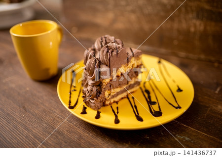 Slice of chocolate layered cake on yellow plate with ceramic mug on wooden table under warm light Slice of chocolate layered cake on yellow plate with ceramic mug on wooden table under warm light 141436737
