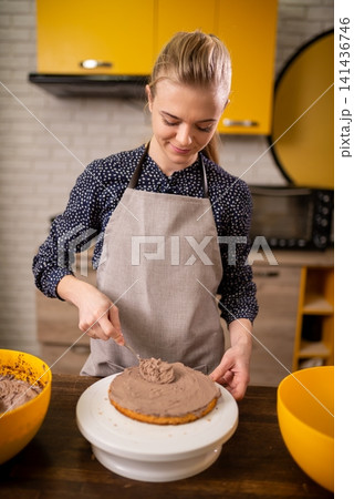 Smiling woman in apron frosting round chocolate cake on turntable in warm modern kitchen Smiling woman in apron frosting round chocolate cake on turntable in warm modern kitchen 141436746