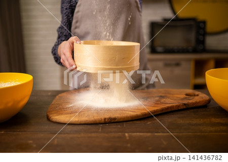 Baker sifting flour over wooden board in warm kitchen scene with ceramic bowls and apron Baker sifting flour over wooden board in warm kitchen scene with ceramic bowls and apron 141436782