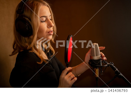 Young woman singing into studio microphone with headphones in warm acoustic booth Young woman singing into studio microphone with headphones in warm acoustic booth 141436793
