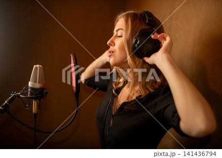 Woman singing into studio microphone with headphones in warm acoustic booth Woman singing into studio microphone with headphones in warm acoustic booth 141436794