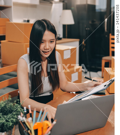 Startup SME small business entrepreneur of freelance Asian woman wearing apron using laptop and box to receive and review orders online to prepare to pack sell to customers, online sme business ideas. Startup SME small business entrepreneur of freelance Asian woman wearing apron using laptop and box to receive and review orders online to prepare to pack sell to customers, online sme business ideas. 141437208