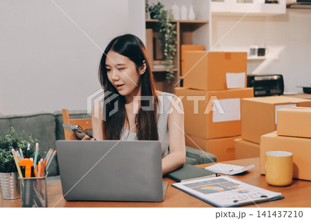 Startup SME small business entrepreneur of freelance Asian woman wearing apron using laptop and box to receive and review orders online to prepare to pack sell to customers, online sme business ideas. Startup SME small business entrepreneur of freelance Asian woman wearing apron using laptop and box to receive and review orders online to prepare to pack sell to customers, online sme business ideas. 141437210