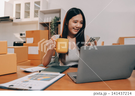 Startup SME small business entrepreneur of freelance Asian woman wearing apron using laptop and box to receive and review orders online to prepare to pack sell to customers, online sme business ideas. Startup SME small business entrepreneur of freelance Asian woman wearing apron using laptop and box to receive and review orders online to prepare to pack sell to customers, online sme business ideas. 141437214